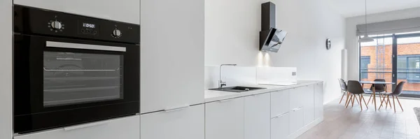 Panorama of long white kitchen with wooden floor and with black sink, oven and kitchen hood, and dining table next to window