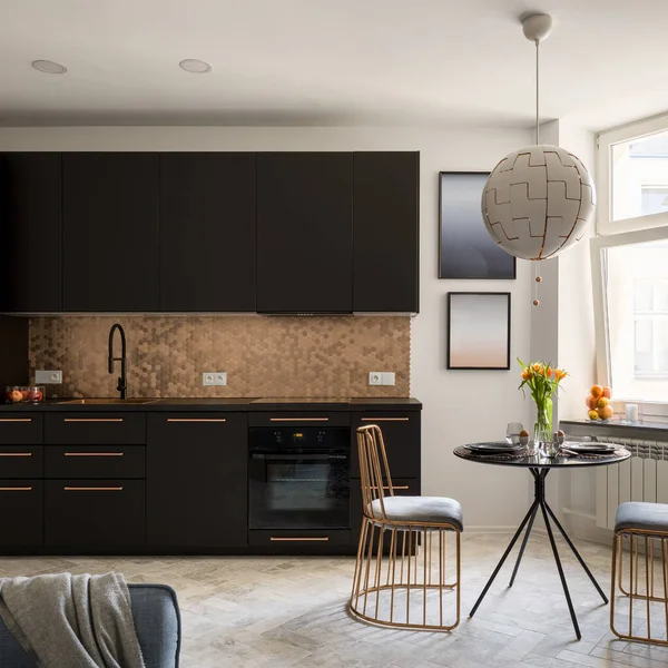 Stylish and small kitchen with black furniture and copper hexagon tiles and with black, dining table and two copper chairs under big ceiling lamp