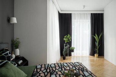 Rahat ve şık bir yatak odası, büyük yataklı, dekorasyonlu siyah komodinli, güzel ahşap zemin ve uzun perdelerin arkasında büyük pencere duvarları.