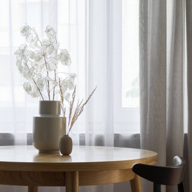 Dekoratif vazolar, beyaz ve bej perdeli pencerenin yanındaki ahşap yemek masasında doğal dekoru olan vazolar.