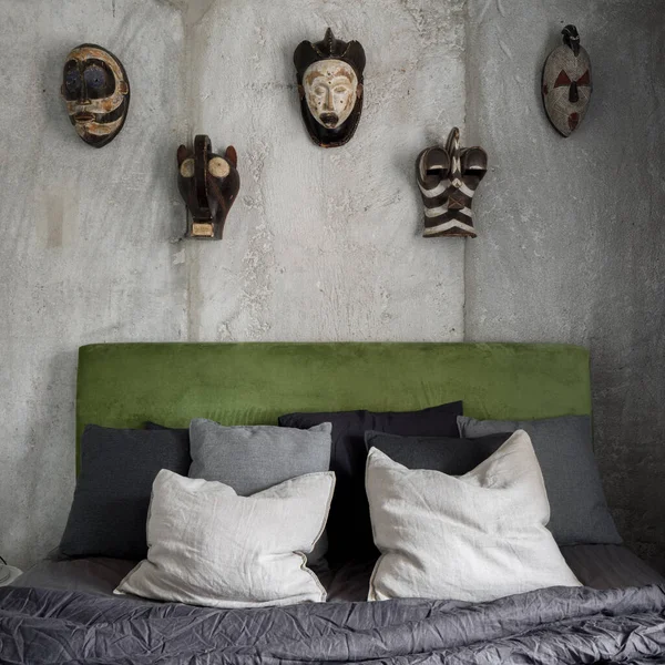 Bare concrete and african masks on wall behind bed with green velour headboard