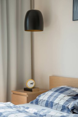 Parlak yatak odasında, üzerinde saat ve kitap olan ahşap komodinin üzerinde siyah kolye lambası.