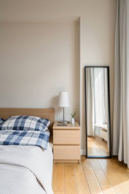 Yatağın yanında lamba ve siyah çerçeveli uzun ayna olan basit ahşap bir komodin.