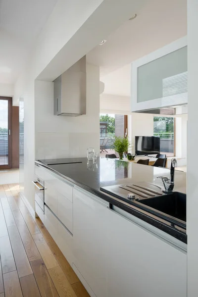 Kitchen area with white walls and cupboards, wooden floor and black countertop open to living room with television screen