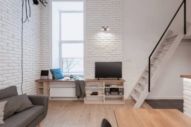 Loft style apartment with stairs to mezzanine from tv room with narrow window and white brick on the walls clipart