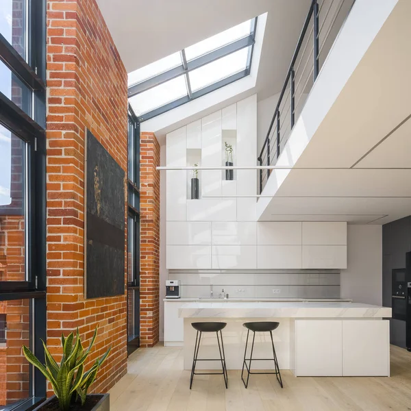 Amazing big windows in loft style apartment with white, spacious and luxury kitchen, exposed red bricks on the walls and mezzanine