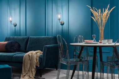 Zarif koltuğu olan şık oturma odasında hoş bir ışık. Pervazlı mavi duvarlar. Şeffaf sandalyeler ve dekorasyonlarla modern yemek masası.