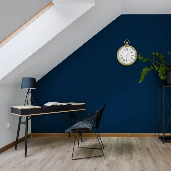 Stylish attic home office room with simple desk and chair and big golden clock on dark blue wall