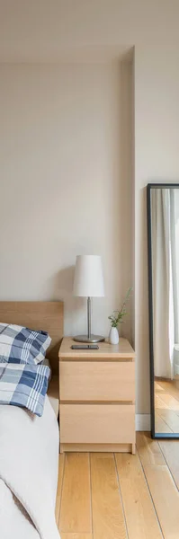 Vertical panorama of simple wooden bedside table with lamp next to bed and long mirror in black frame