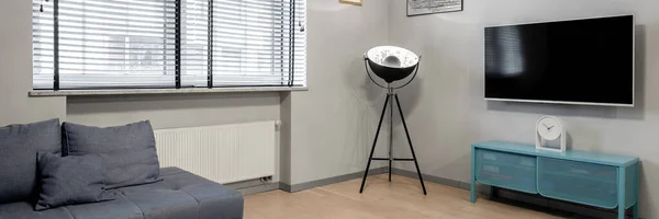 Panorama of small living room with decorative, black lamp on tripod, television screen, blue sideboard, gray sofa and with big window with blinds