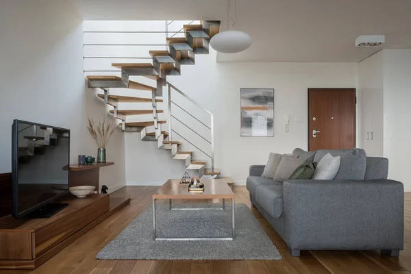 Simple living room with comfortable couch, wooden coffee table and television screen on wooden sideboard and with modern stairs to second floor