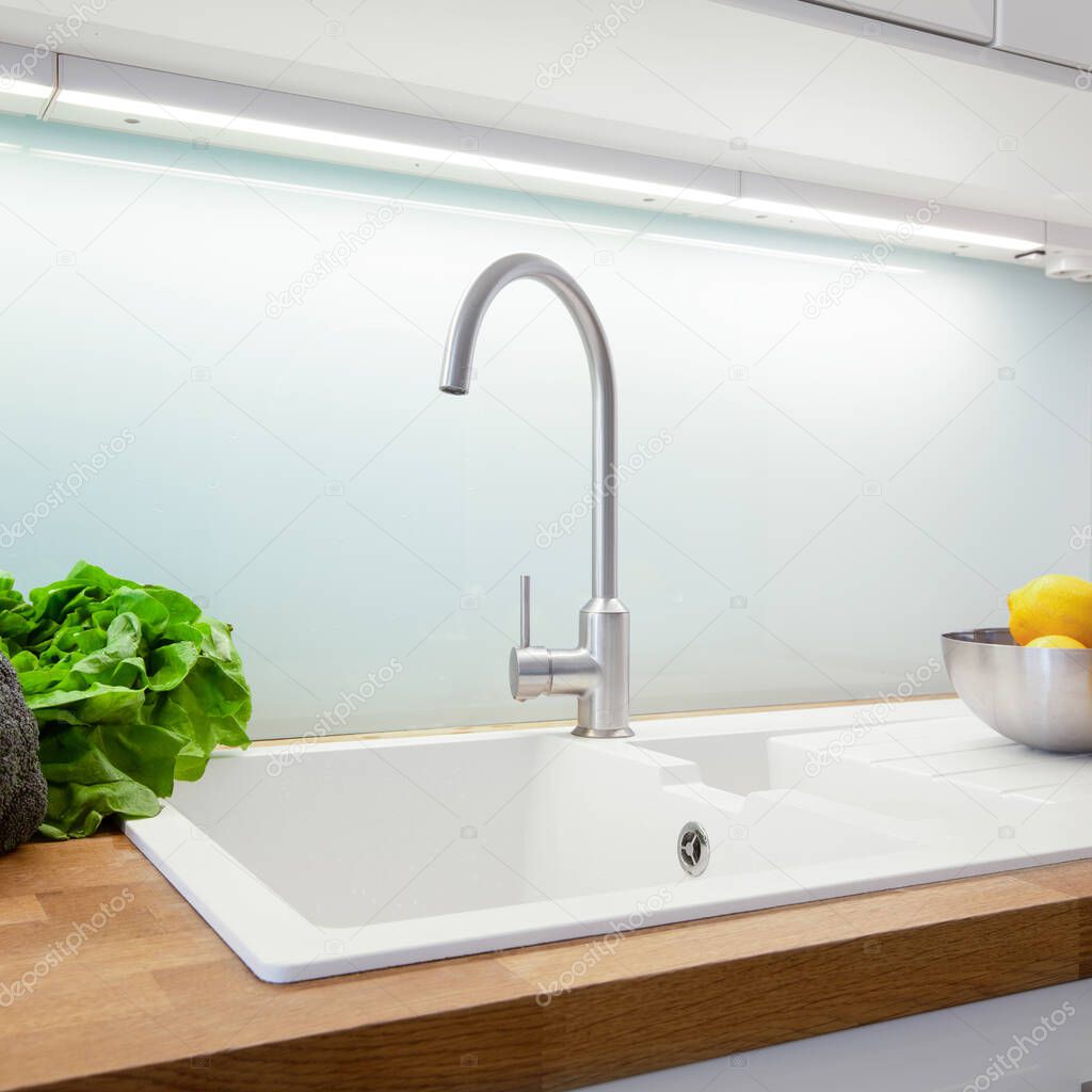 Close-up on simple, white kitchen sink with silver faucet, glass backsplash, led light and wooden countertop