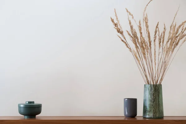 Stylish green container, simple blue vase and modern, glass, green vase with dried grass on wooden shelf with beige wall in background