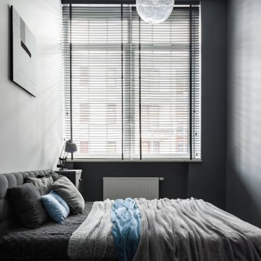 Büyük ve rahat yataklı gri renkli küçük ve şık bir yatak odası ve siyah panjurlu büyük bir pencere.