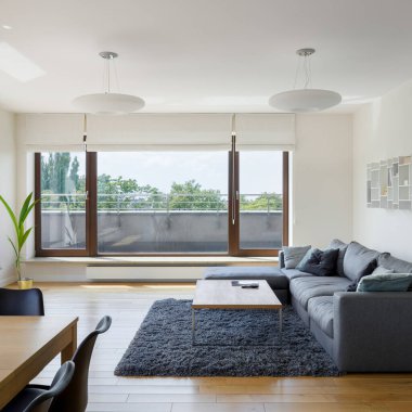 Geniş ve parlak oturma odası, basit mobilyalar ve terasa açılan büyük pencere kapıları.