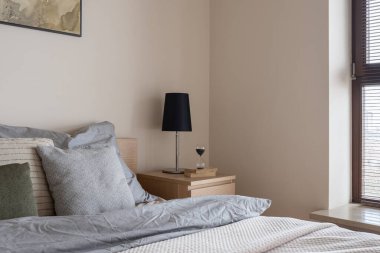 Küçük yatak odasındaki rahat yatağın yanında kitap ve kum saati olan ahşap komodinin üzerinde siyah lamba.