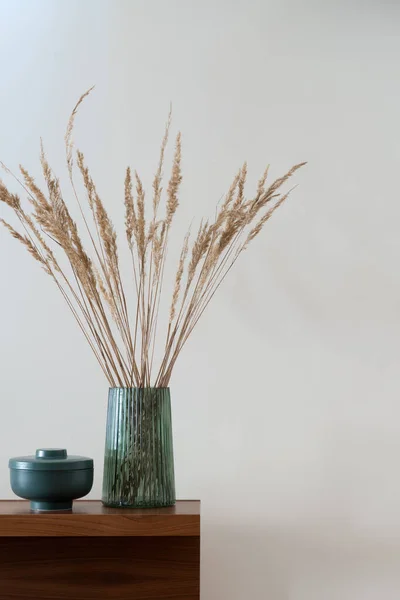 Decorative dried grass in stylish, glass, green vase next to nice green container on wooden sideboard, close-up