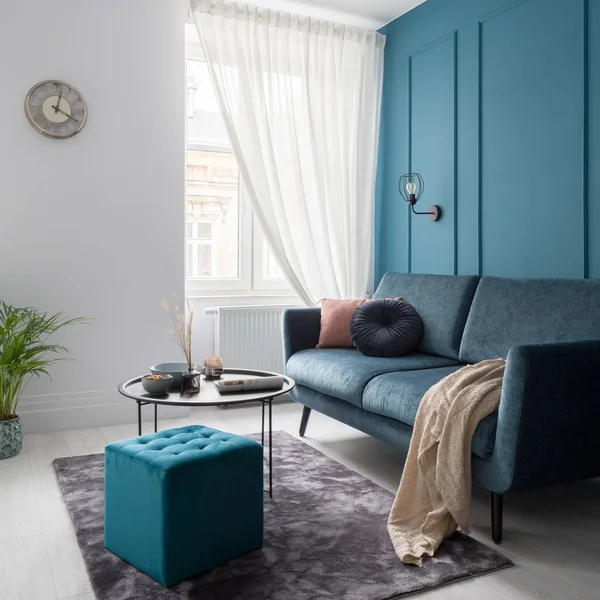 Small living room with teal blue wall with decorative molding, modern sofa, black coffee table and square, quilted ottoman