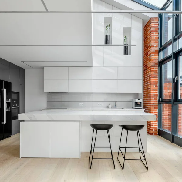 Modern designed kitchen in loft style apartment with white furniture, brick wall and big window from ceiling to floor