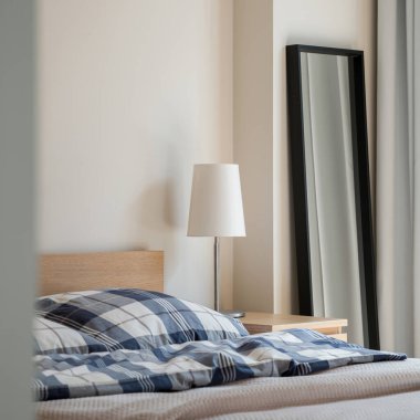 Basit, beyaz lamba, yatağın yanındaki ahşap komodinin üzerinde ve siyah çerçevede ayna.