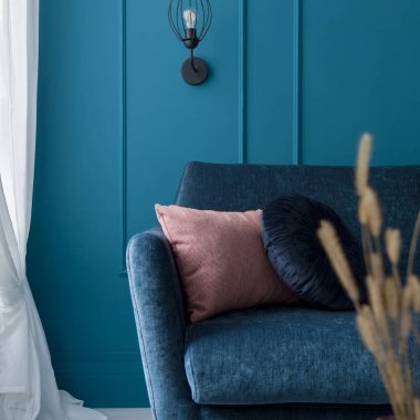 Odada dekoratif yastıklar olan mavi bir kanepeye yakın plan. Küflenmiş mavi duvar, beyaz perdeler ve siyah lamba.