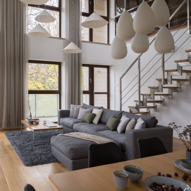 İki katlı lüks bir daire, büyük pencereli, geniş oturma odası rahat, gri köşeli kanepe ve merdivenli.