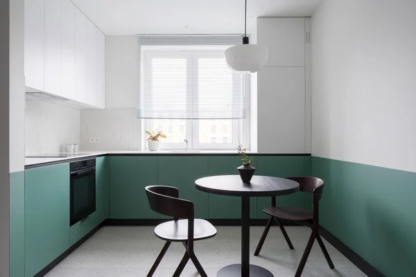 Simple and modern design in kitchen with window, half green and white walls and cupboards and with black table with two chairs