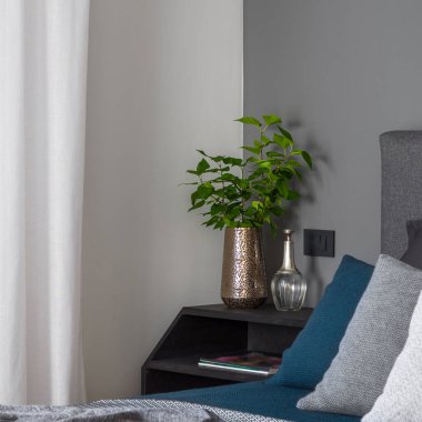 Close-up on stylish golden vase with green plant and decorations on simple, black nightstand next to cozy bed in bright bedroom