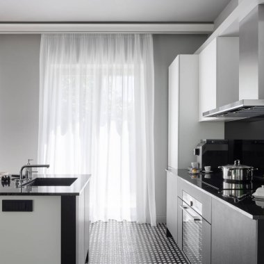 Modern black and white kitchen with big windows behind white curtains and with black and white kitchen island with sink and black and white furniture with black countertops