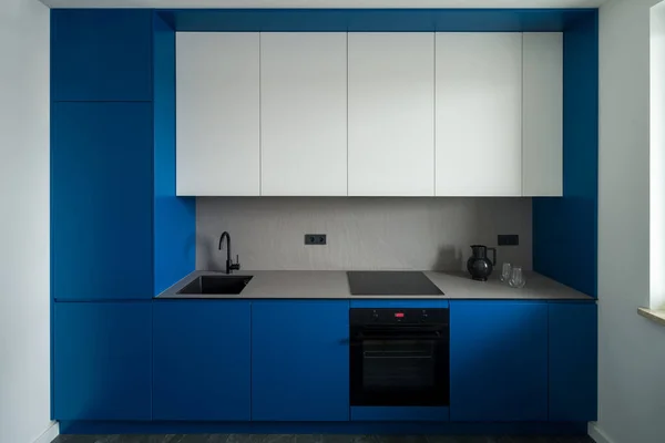 Simple, modern and small kitchen with blue and white cupboards and gray countertop and backsplash and black sink