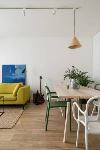 Eclectic style living room with yellow couch, art, wooden floor and ceiling light and with dining area with wooden table and different white and green chairs