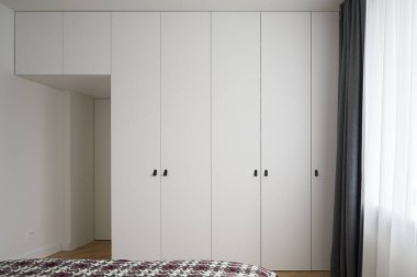 Pencerenin yanında beyaz ve gri perdeleri olan modern, beyaz gardırop duvarlı geniş bir yatak odası.