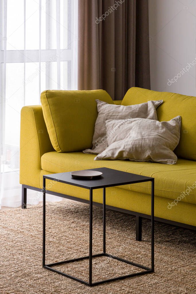 Stylish black coffee table on natural carpet and modern yellow couch with decorative pillows in bright living room