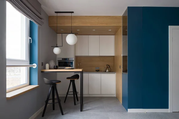 Modern and small kitchen with console table and two chairs, stylish white furniture, wooden walls and backsplash, gray floor tiles, blue wall and window