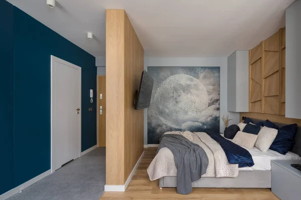 Stylish bedroom with comfortable bed, wooden walls and decorative wall mural next to corridor with blue wall