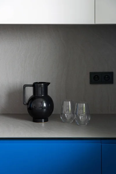 现代灰色厨房台面上有两个玻璃杯的时尚黑色水壶 新厨房里有蓝色和白色的碗橱 — 图库照片