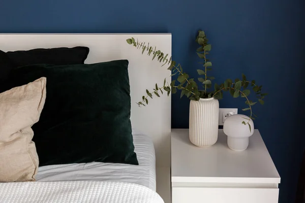 White bedside table with decorative lamp and vase with eucalyptus twigs next to cozy bed in bedroom with navy blue wall