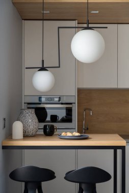 Dekoratif lambalı küçük mutfak, beyaz mobilyalar ve iki sandalyeli ahşap masa.