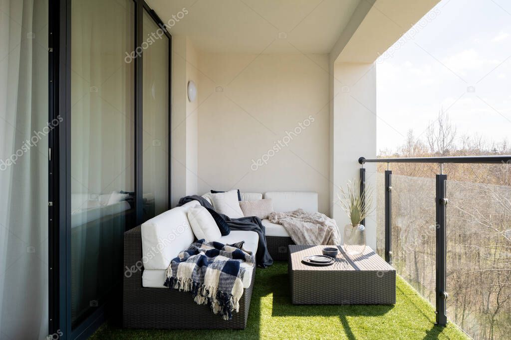 Cozy decorated balcony with big window doors to apartment, synthetic grass and stylish rattan corner sofa with decorative pillows and rattan coffee table