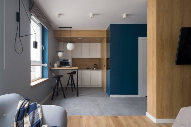 Small and modern kitchen with white cupboards, wooden wall and table open to room with cozy armchair