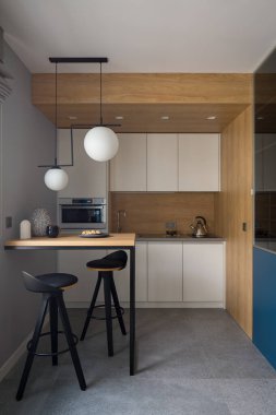 Small and modern kitchen with white furniture, wooden walls and table with two, black chairs and granite floor tile 