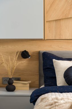 Black vase with dried grass and books on gray bedside table next to comfortable bed with stylish bedclothes