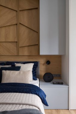 Parlak yatak odasındaki şık dekorasyonlu yatağın yanında gri komodinin yanında, duvarında ahşap süslemeler var.