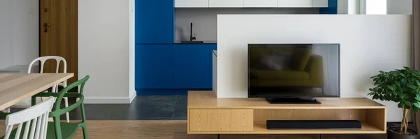 Panorama of simple tv room with wooden sideboard open to dining area with table and eclectic white and green chairs open to modern kitchen with white and blue furniture