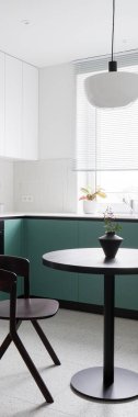 Modern, siyah ve yuvarlak yemek masasının dikey manzarası şık bir lambanın altında büyük pencereli, beyaz ve yeşil dolaplı ve terrazzo zeminli yeni tasarım mutfağında.
