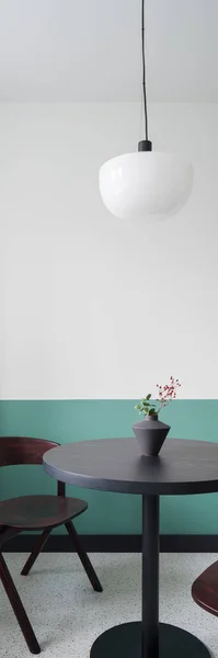 Vertical panorama of stylish, round, wooden dining table under modern lamp in room with decorative white and green wall and terrazzo floor