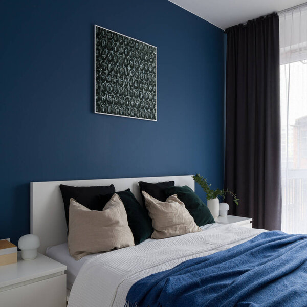 Modern bedroom with white furniture, comfortable bed, window behind curtains and stylish blue wall with art