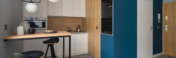 小厨房全景 白色的橱柜 木制的墙壁 装饰灯下的桌子 旁边有蓝色的墙和主入口的走廊 — 图库照片