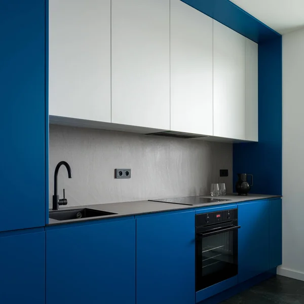 Simple and modern design in kitchen with stylish blue walls and cupboards, gray countertop and backsplash, white upper cabinets and black sink, tap and oven