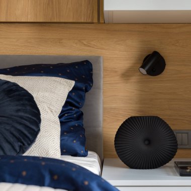 Zarif bir vazoya yakın plan basit bir komodinin üzerinde, ahşap duvarda siyah bir lamba, şık çarşaflı rahat bir yatağın yanında.